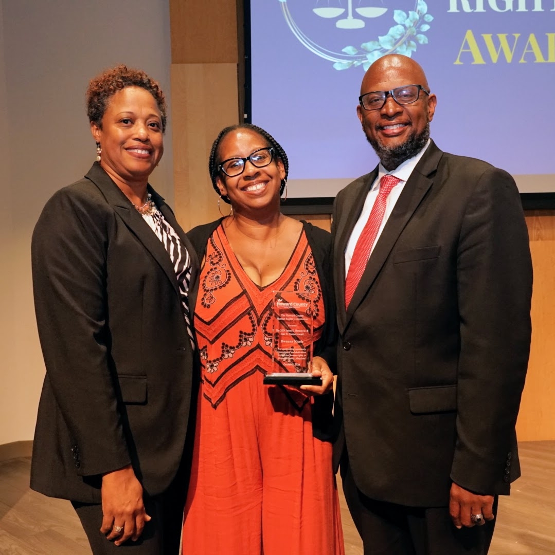 Three people in business formal dress. The person in the center holds a clear glass award. 