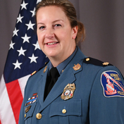 Major Cory Zirk, in uniform, standing in front of an American flag with grey background.