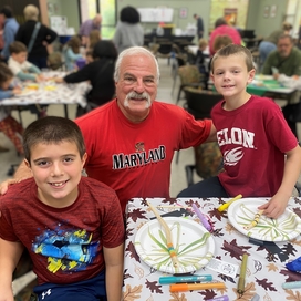 Older adult white male with two kids elementary school age, all wearing red t-shirts posing together smiling at an arts & crafts table.